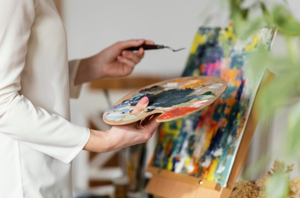 woman in white clothing painting with acrylics and holding a palette