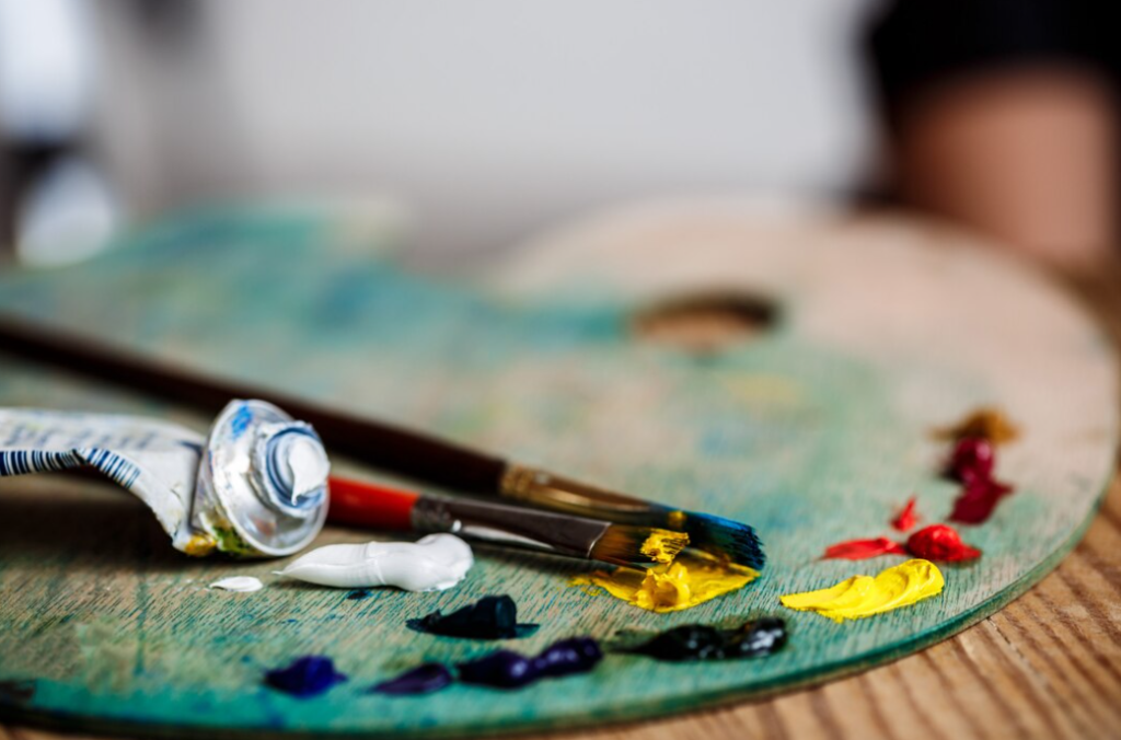 An artist's palette with vibrant blobs of paint and a brush, with a paint tube